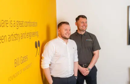 Two IT Support workers stood infront of a yellow wall smiling.