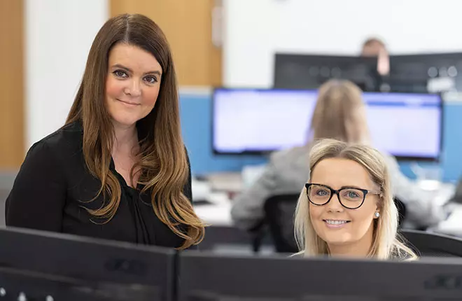 Two women in business attire behind computer smiling at camera