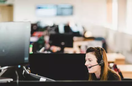 Woman wearing telephone headset sat in an open plan office